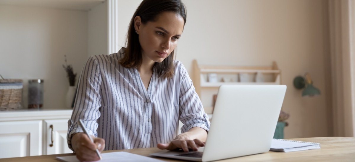 white brunette woman looking at laptop and making notes with a pen on paper